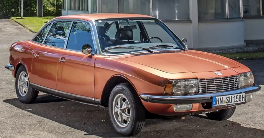 A vintage orange four-door car with chrome accents is parked on a street in front of a building with large windows. The car has a European license plate and a classic 1970s design.