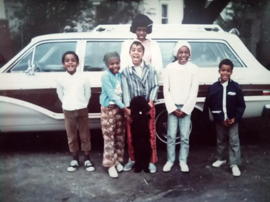 Six children and an adult stand smiling in front of a vintage white station wagon with wood paneling, on a street with trees and houses in the background.