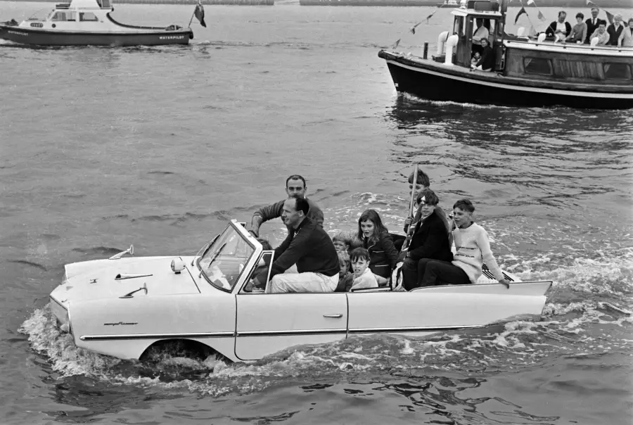 A group of people ride in a white amphibious car driving through water, with boats in the background. The scene appears cheerful and lively.