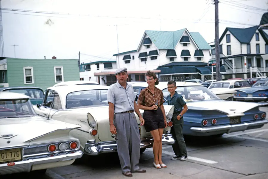 A man, woman, and boy stand by classic cars in a parking lot, with vintage houses in the background on a sunny day.