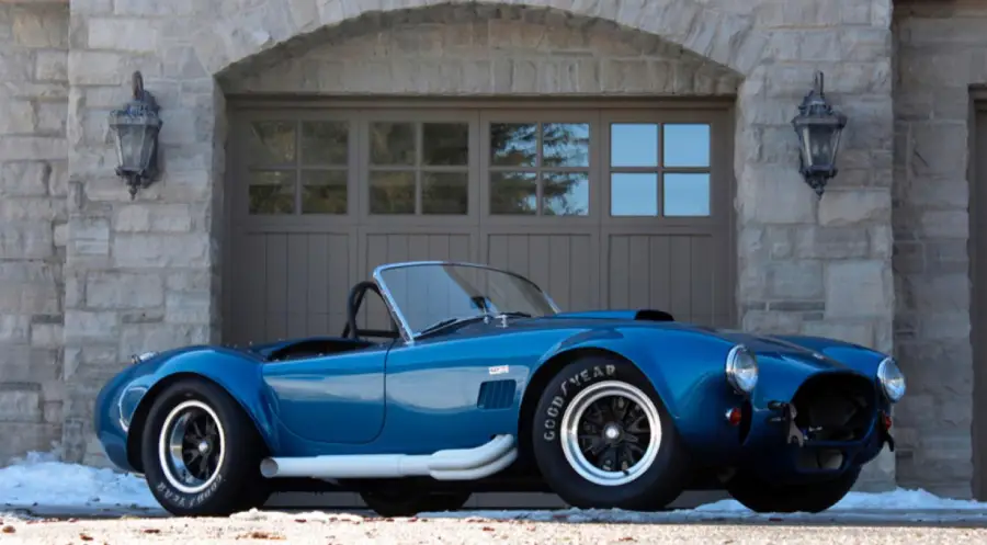 A blue classic Shelby Cobra convertible sports car is parked in front of a stone garage with two wall lanterns and closed double doors.