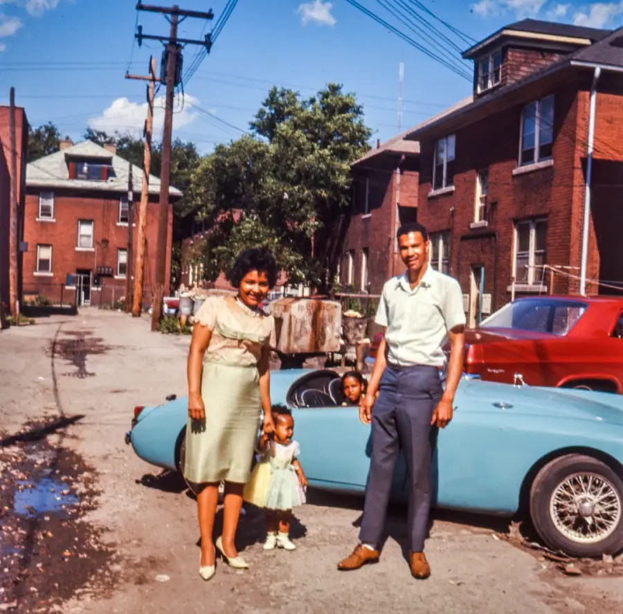 A family of four stands in front of a blue convertible car in an urban alley, surrounded by brick buildings and a red car. The parents and two children are dressed in vintage clothing, smiling on a sunny day.