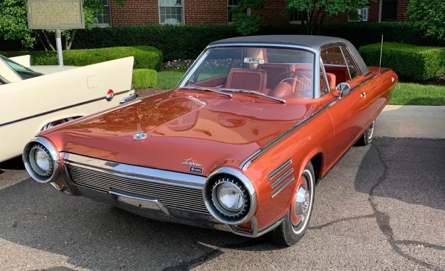 A classic two-door car with a red exterior, chrome trim, and a black roof is parked on a street. The car has a unique round headlight design and a vintage license plate. Shrubs and a brick building are in the background.