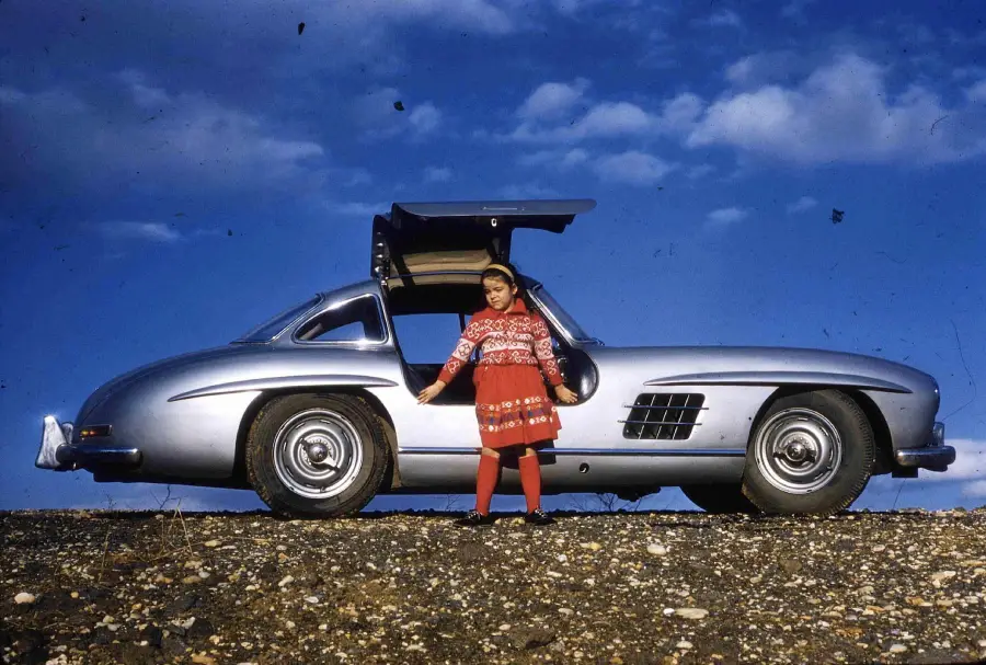 A child in a red outfit stands next to a silver classic car with its gullwing door open, set against a blue sky with scattered clouds and standing on rocky ground.