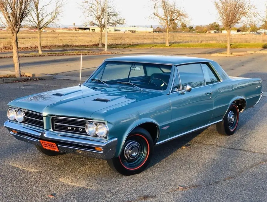 A light blue 1964 Pontiac GTO classic car parked in an empty lot on a sunny day, with bare trees and open fields in the background.