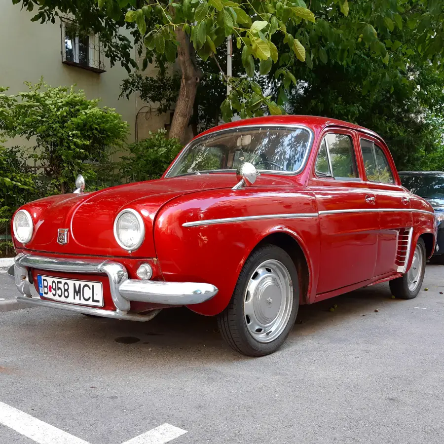 A shiny red vintage car is parked on a street beside trees and a building, with a Romanian license plate visible on the front bumper.