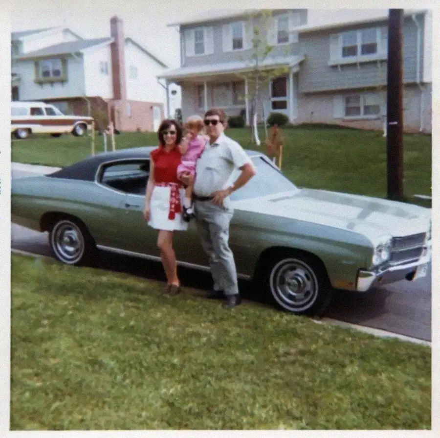 A woman, man, and child pose in front of a green classic car parked on a suburban street, with houses and a station wagon visible in the background.