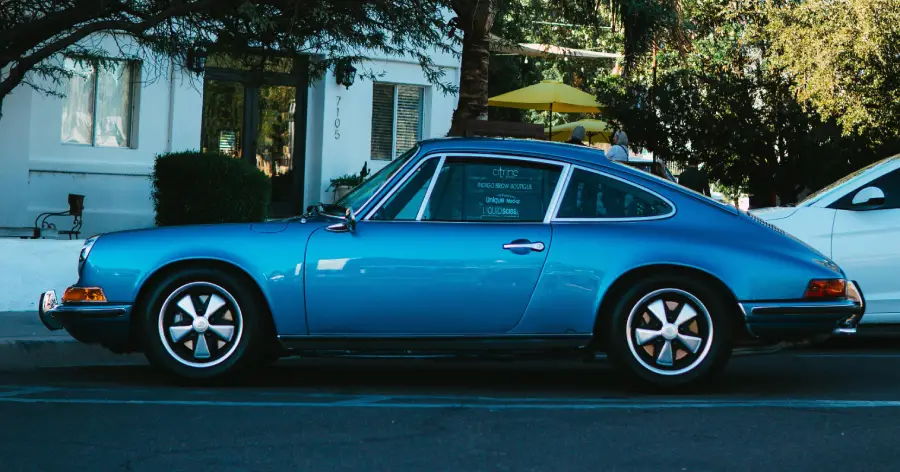 A classic blue Porsche sports car is parked on the street in front of a white building and some trees, with sunlight casting shadows.