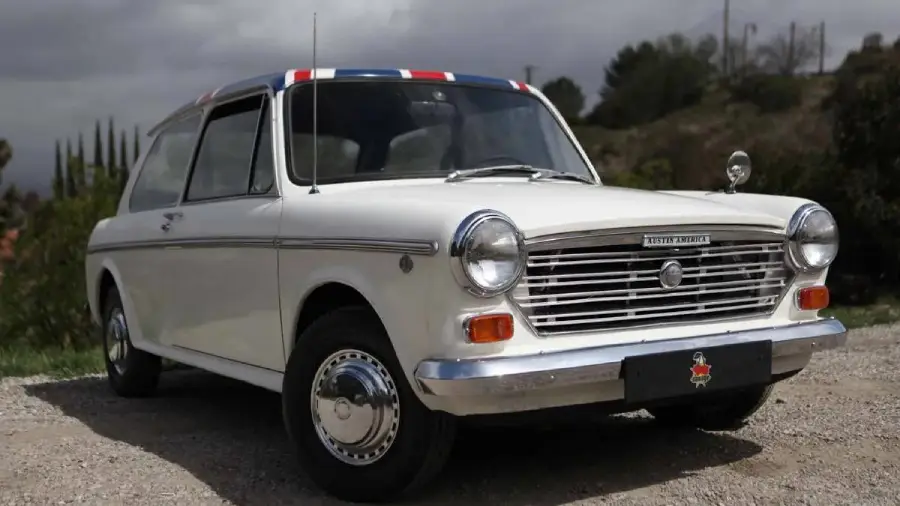 A white classic Austin America car is parked outdoors on a gravel surface, with trees and a cloudy sky in the background. The roof edge is decorated with red, white, and blue stripes.