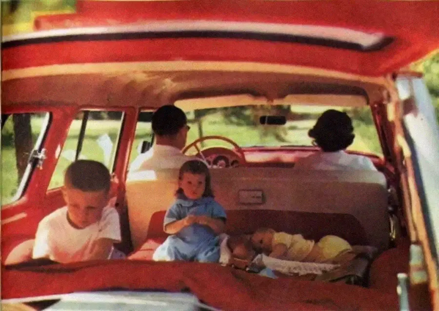 Two adults sit in the front seats of a red car, while three children are in the back: one boy sits upright, a toddler sits in the middle, and a baby lies down, with sunlight streaming in through the windows.