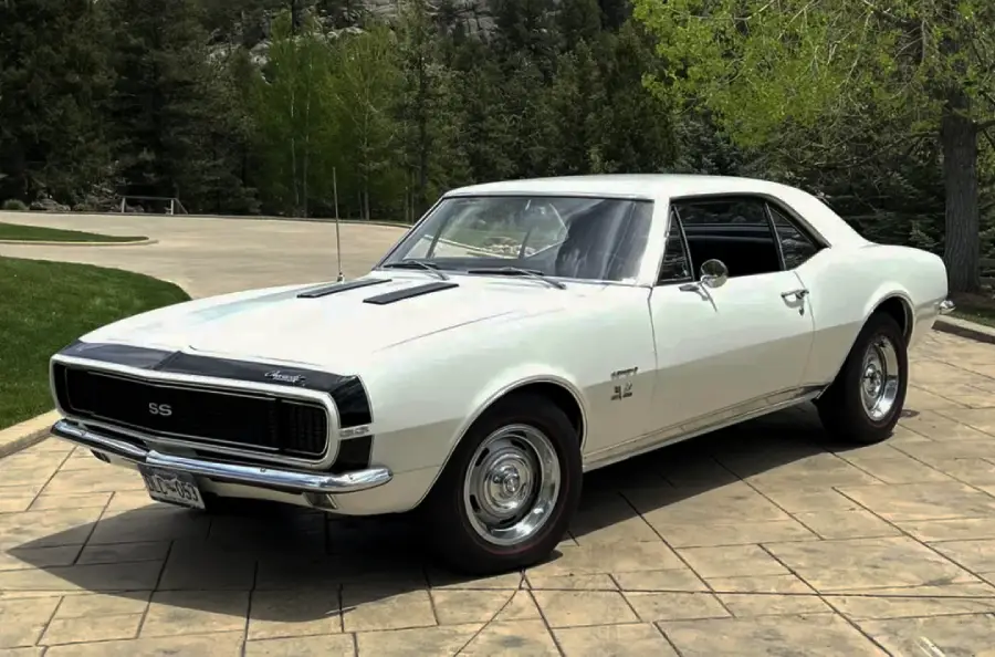 A classic white 1967 Chevrolet Camaro SS coupe is parked on a stone driveway, surrounded by greenery and trees under a partly cloudy sky.