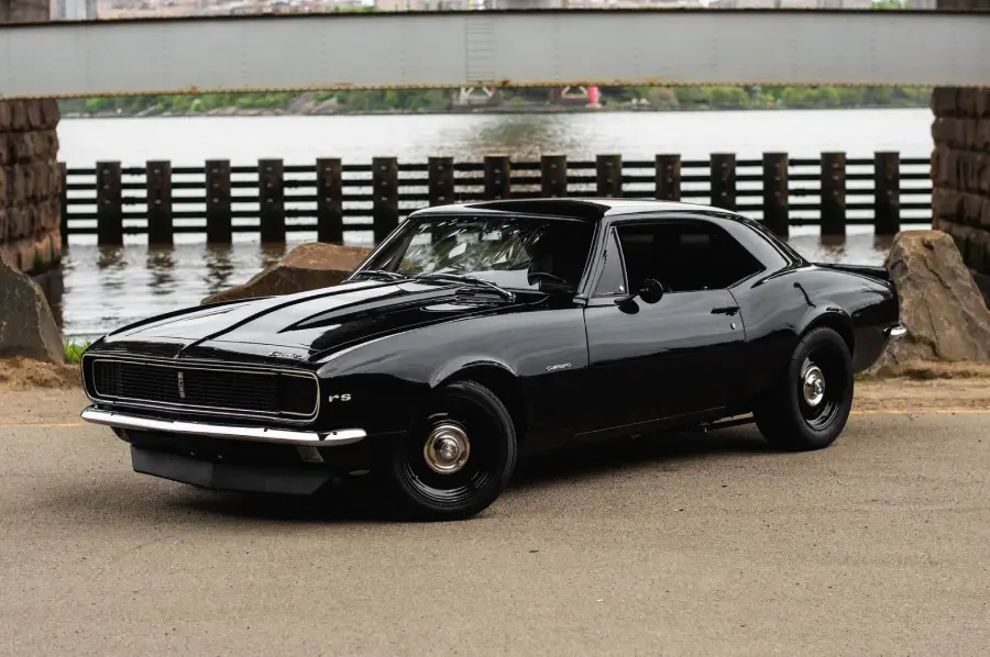 A black classic muscle car, likely a late 1960s Chevrolet Camaro, is parked on a paved road near a body of water, with a bridge and rocks in the background.
