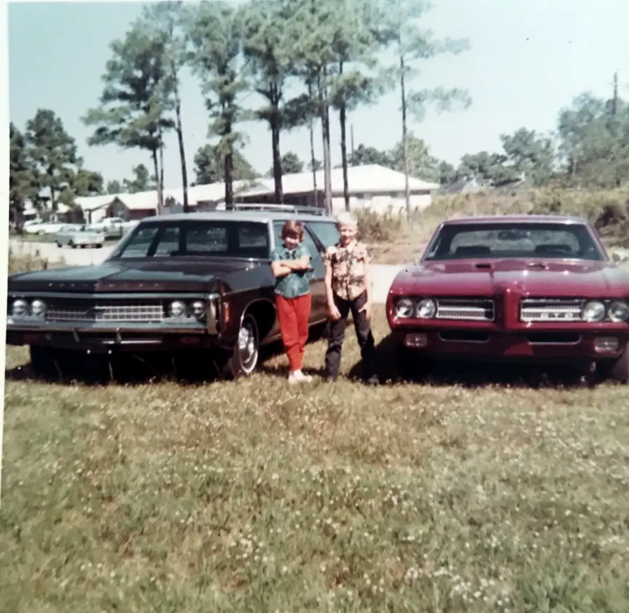 Two people stand between a black car and a red car on grass, with trees and houses in the background, on a sunny day.