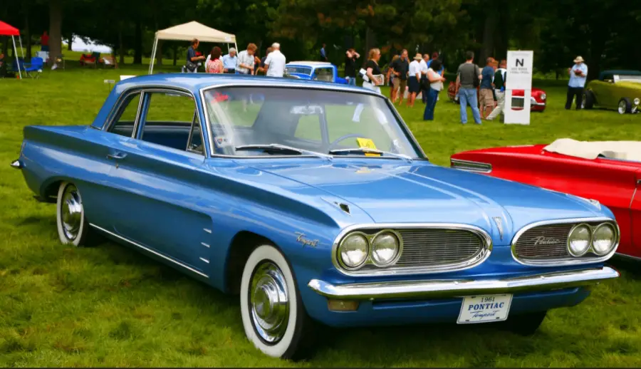 A light blue 1962 Pontiac Tempest coupe is parked on grass at a classic car show, with people and other vintage vehicles visible in the background.