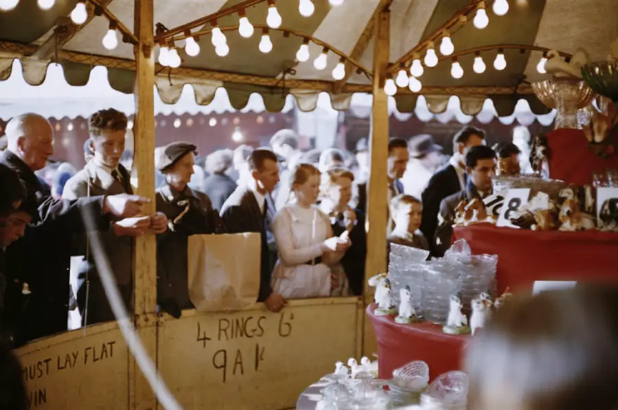 People stand under a striped tent at a fair, watching a game stall filled with ceramic prizes and lightbulbs, while a man appears to organize or announce something. Signs and paper bags are visible.
