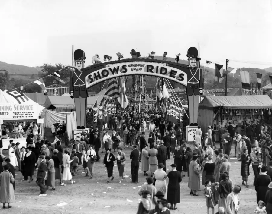 Black-and-white photo of a crowded fairground with people walking under an archway labeled “Shows and Rides,” surrounded by flags, tents, and attractions. The scene is lively and festive.