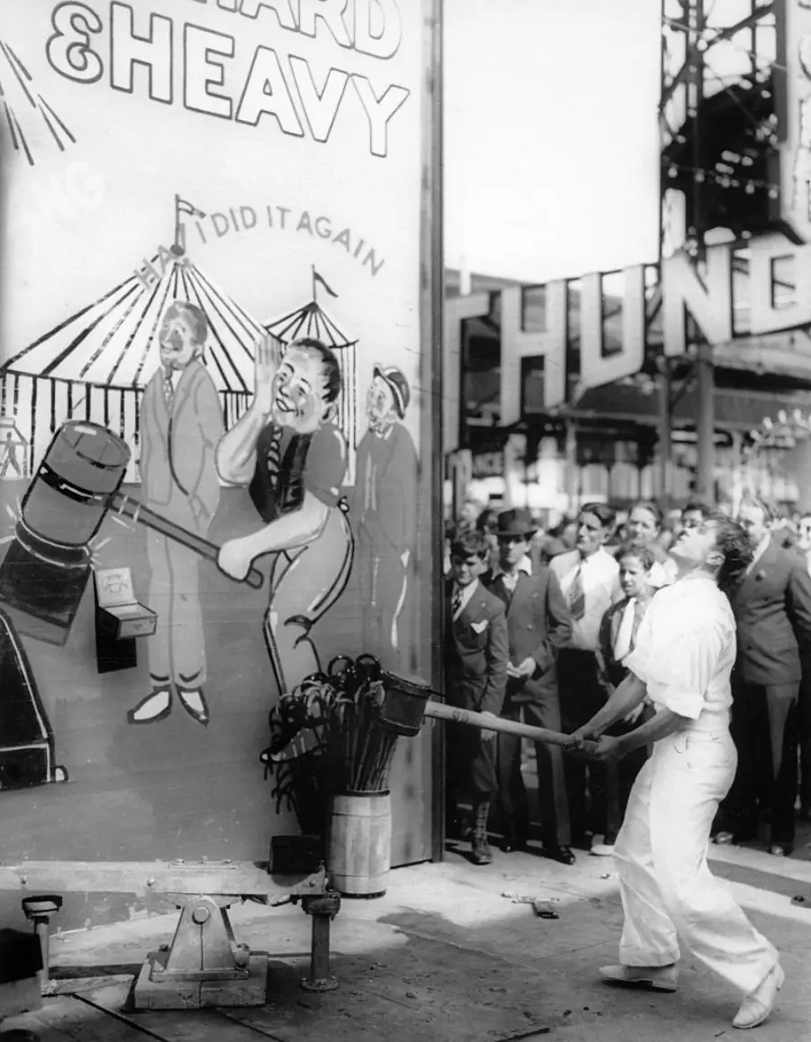 A person swings a large mallet at a carnival strength tester while a crowd watches. A painted sign behind them shows a circus scene and reads “HARD & HEAVY.”