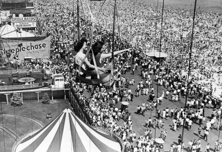 Two women ride a swing ride above a crowded boardwalk and beach. One points at something in the distance. A “Steeplechase” sign and tent roof are visible among the dense crowd below on a sunny day.