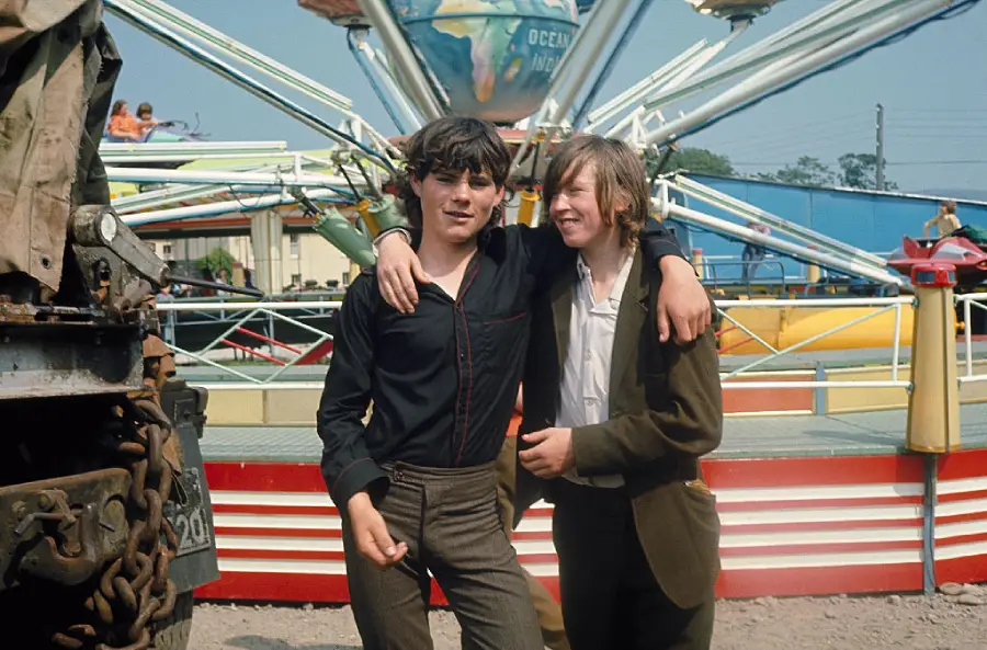 Two boys stand arm-in-arm, smiling, in front of a colorful amusement park ride with a globe at the top. One wears a black shirt and striped pants; the other wears a brown jacket. The scene is outdoors, on a sunny day.