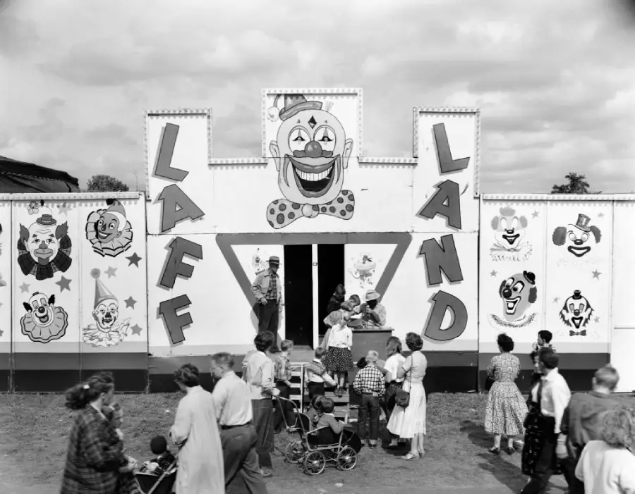 Black-and-white photo of families and children at a carnival, standing outside a funhouse called “Laff Land” decorated with large clown faces and letters; people with strollers gather near the entrance.