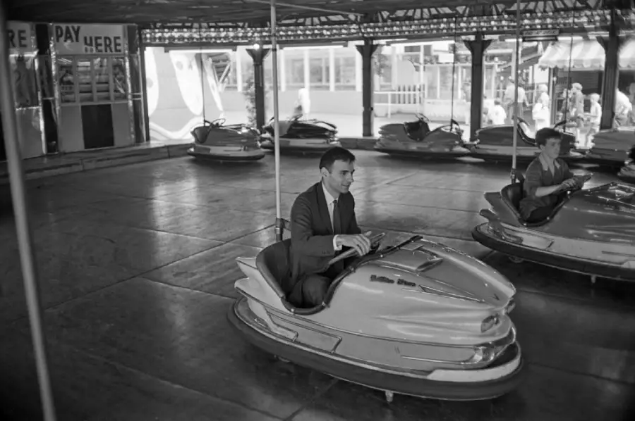A man in a suit rides a bumper car indoors at an amusement park, while another person drives a bumper car nearby. Several empty bumper cars are visible in the background.