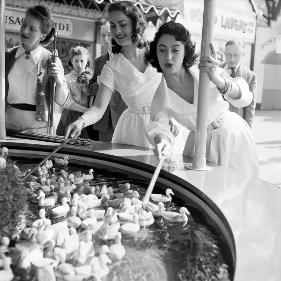 Women in vintage dresses play a hook-a-duck game at a fair, using sticks to catch floating toy ducks in a water trough while onlookers watch. The scene appears lively and joyful, with bright expressions on their faces.