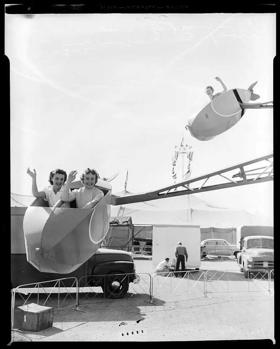 Three people ride a spinning carnival ride, smiling and waving. Two women share one car in the foreground, while another person rides alone in a car above. Cars and tents are visible in the background.