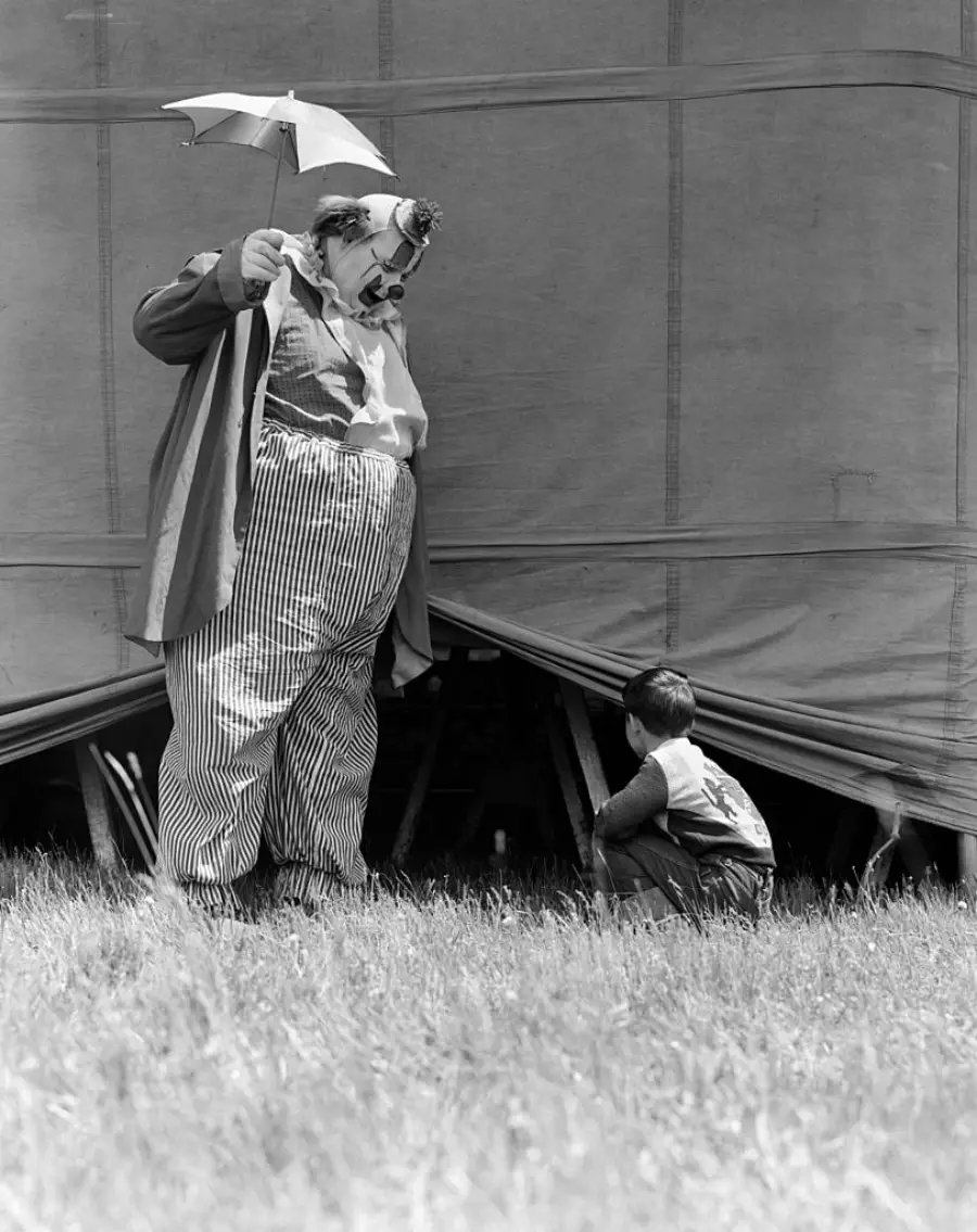 A clown holding a small umbrella stands near a tent, looking down at a young boy who is crouched by the tent’s entrance on a grassy field.