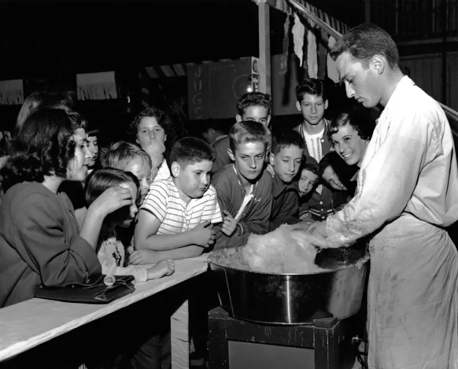 A man makes cotton candy as a group of children and teenagers watch eagerly across a counter at what appears to be a fair or carnival, all in black and white.