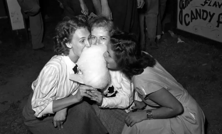 Three women sit close together, laughing and sharing a large piece of cotton candy at an outdoor event, likely a fair or carnival. They all wear vintage clothing, and a candy stand sign is visible in the background.