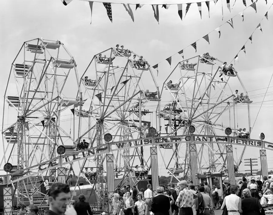 Black and white photo of a crowded fairground with three Ferris wheels, people riding on them, and many fairgoers walking below. Bunting flags hang overhead.