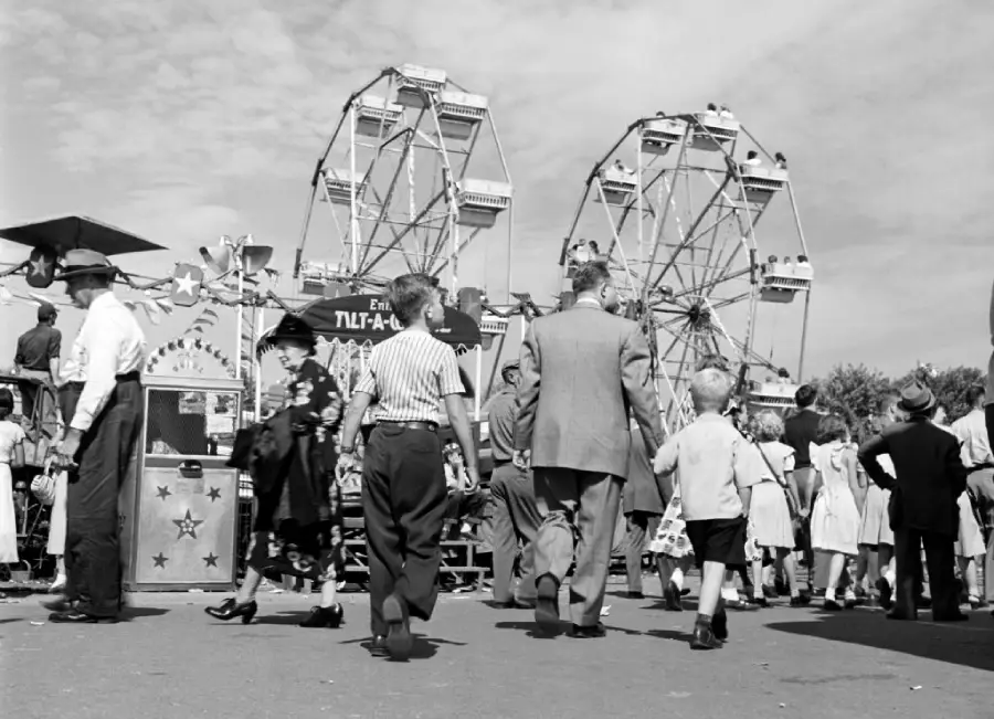 Black and white photo of people at a fair, with two Ferris wheels in the background. Adults and children walk toward the rides, and banners and booths line the crowded scene.