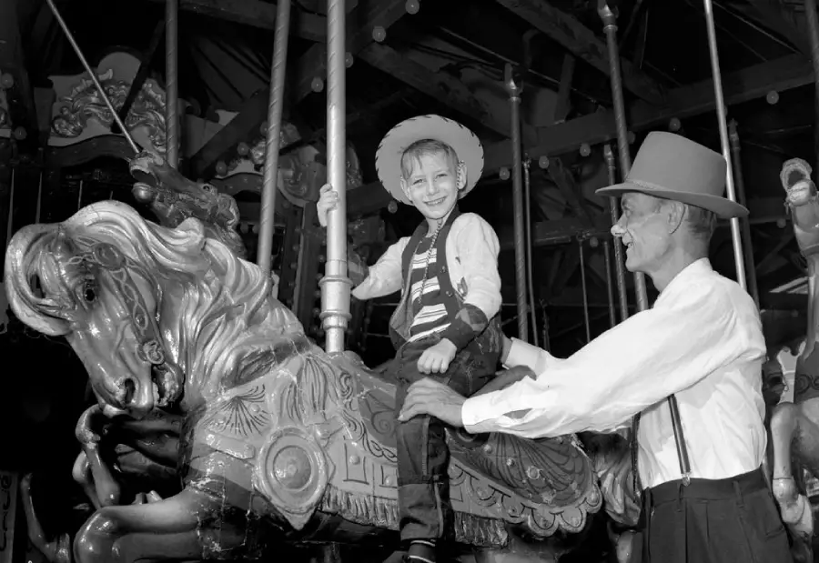 A smiling child in a cowboy hat rides a decorative carousel horse while an older man in a hat steadies him, both enjoying the moment at an amusement park.