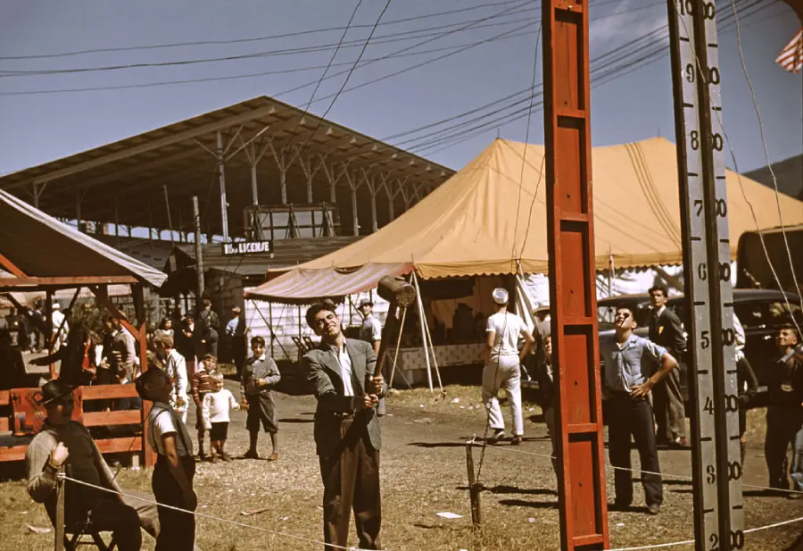 A man swings a mallet at a high striker game at an outdoor fair, surrounded by children and adults. Tents and a grandstand are visible in the background under a sunny sky.
