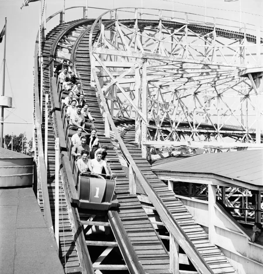 People ride a wooden roller coaster, descending a steep track with hands up. The coaster is marked with the number 1. The structure is large and intricate, with beams and supports visible in the background.