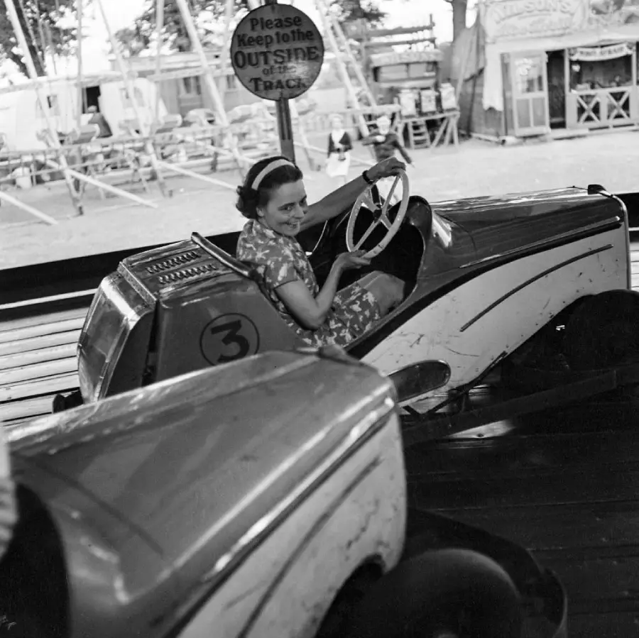 A smiling woman in a patterned dress drives a small ride car at an amusement park. A sign above says, “Please keep to the outside of the track.” Other rides and booths are visible in the background.
