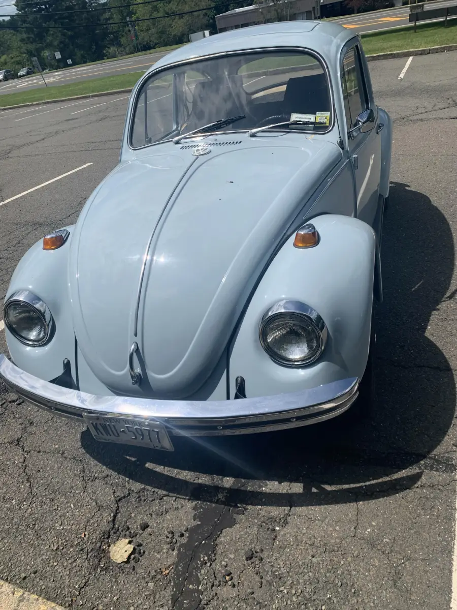A light blue classic Volkswagen Beetle is parked in an empty outdoor parking lot on a sunny day. The car’s front end is facing the camera.