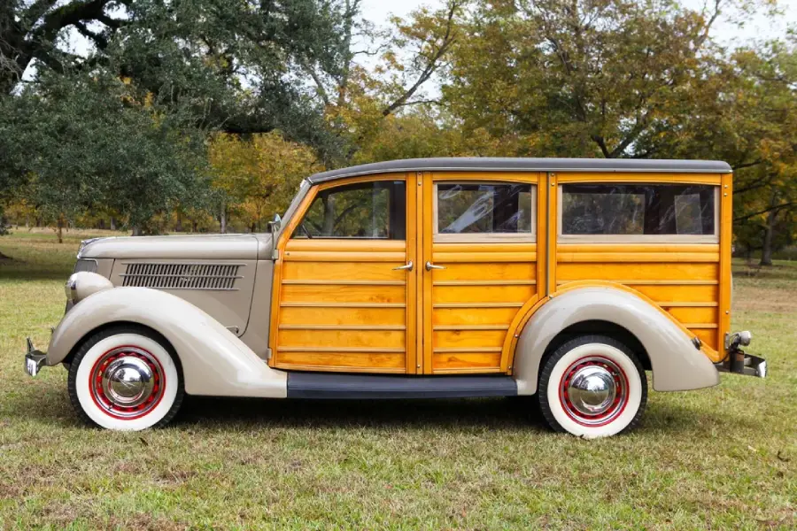 Vintage beige and wood-paneled station wagon parked on grass, featuring whitewall tires and red-rimmed wheels, with trees and a cloudy sky in the background.