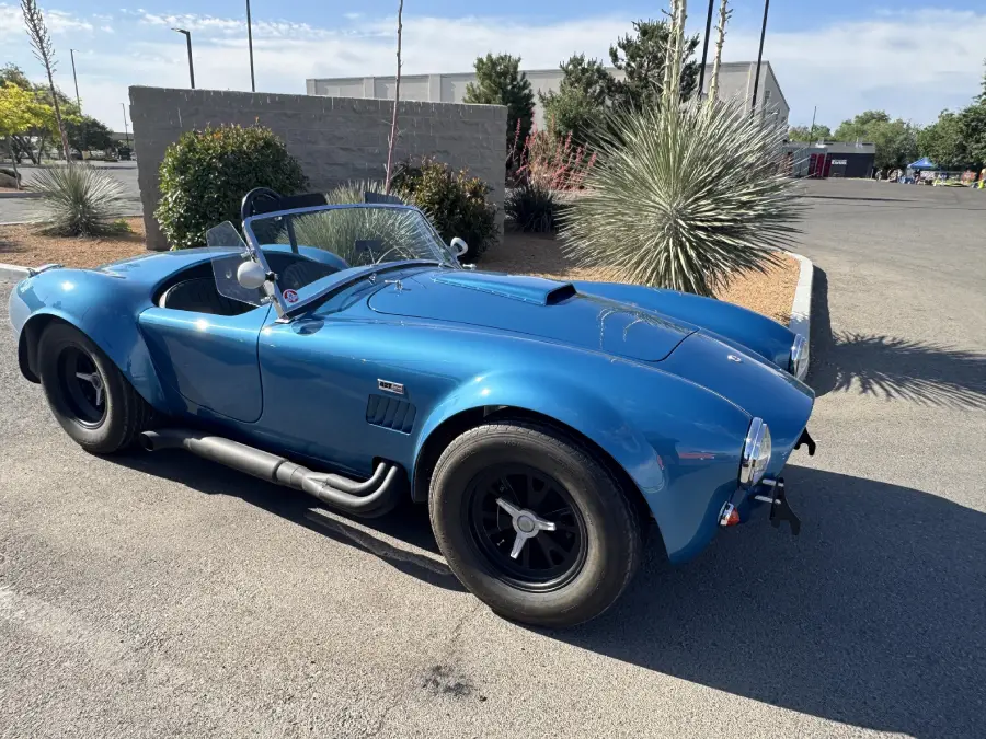 A blue classic convertible sports car with black wheels is parked on a street near desert landscaping and a low concrete wall.