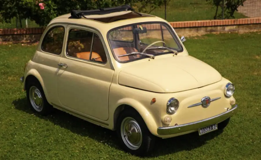 A vintage yellow Fiat 500 with a black convertible roof is parked on grass near a brick border and garden.
