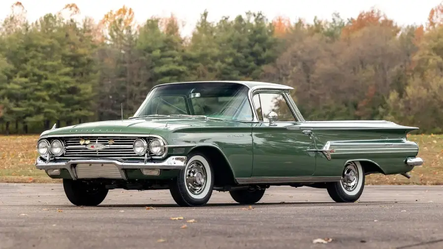 A vintage green Chevrolet El Camino parked on pavement, with trees in the background displaying early autumn foliage.