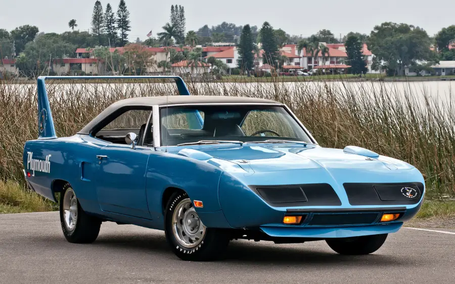 A blue classic Plymouth muscle car with a large rear wing is parked on a road near tall grass, with water and houses visible in the background.