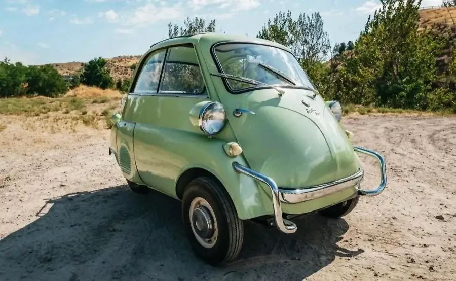 A vintage light green microcar with a bubble-like shape is parked outdoors on a dirt path, surrounded by trees and hills under a partly cloudy sky.