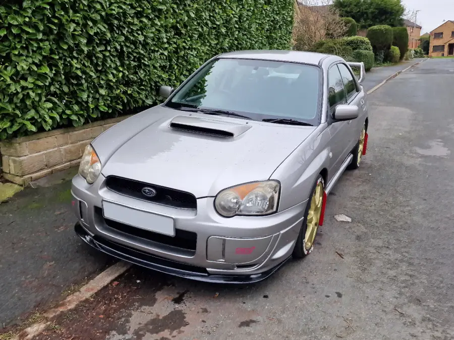 A silver Subaru Impreza with gold wheels and a hood scoop is parked on a residential street next to a tall green hedge.
