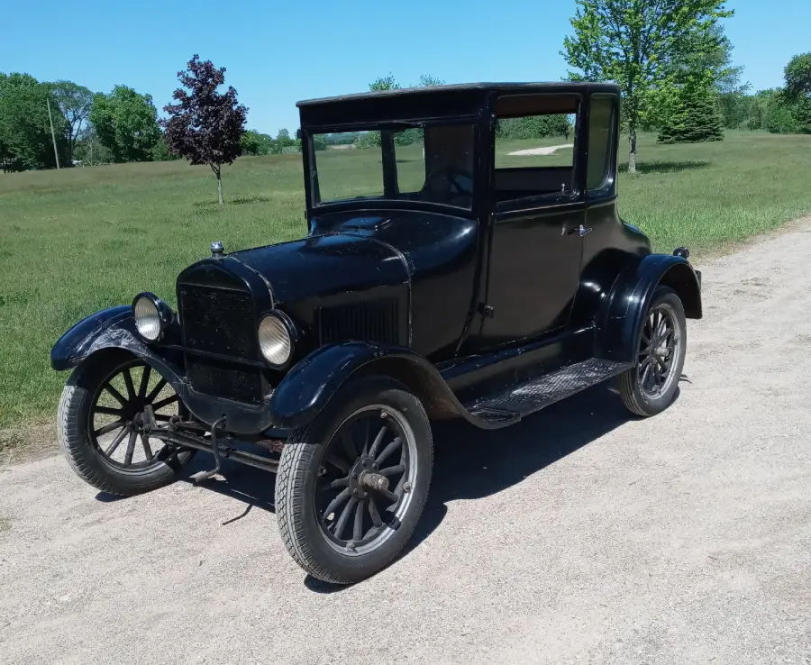 A classic black vintage car is parked on a dirt road in a grassy, open area with trees in the background under a clear blue sky.