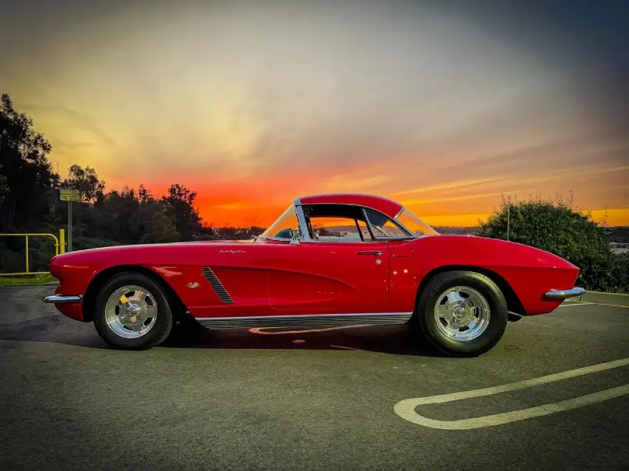 A classic red sports car is parked on pavement at sunset, with a vibrant orange and yellow sky in the background and some trees and bushes nearby.