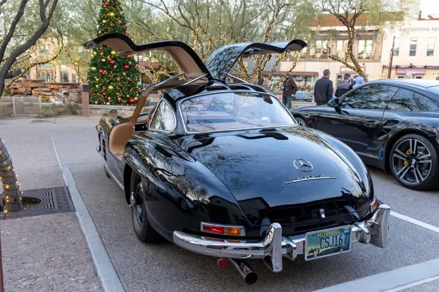 A black classic Mercedes-Benz 300SL with gullwing doors open is parked outdoors near a decorated Christmas tree and another car. People and buildings are in the background.