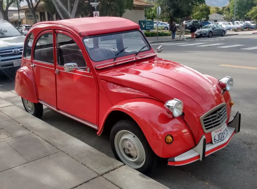 A red vintage Citroën 2CV car is parked on a street by the sidewalk, with trees, parked cars, and buildings visible in the background.