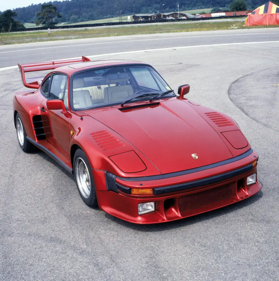 A red Porsche 911 sports car with wide fenders and a large rear spoiler is parked on a paved area near an airstrip, with grassy fields and trees in the background.