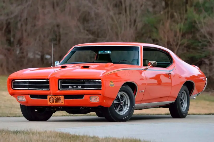 A classic orange 1969 Pontiac GTO muscle car parked on a road, featuring a “GR-RRR!” license plate, hood scoops, black wheels, and chrome trim, with trees and grass in the background.